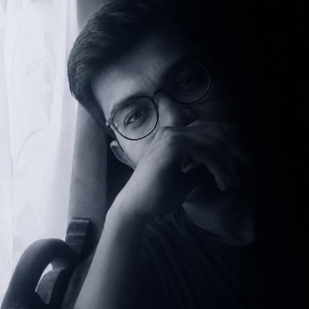 Black-and-white portrait of Raunak Karn, Urdu-Hindi ghazal poet, wearing glasses and resting his chin on his hand beside a sunlit curtain